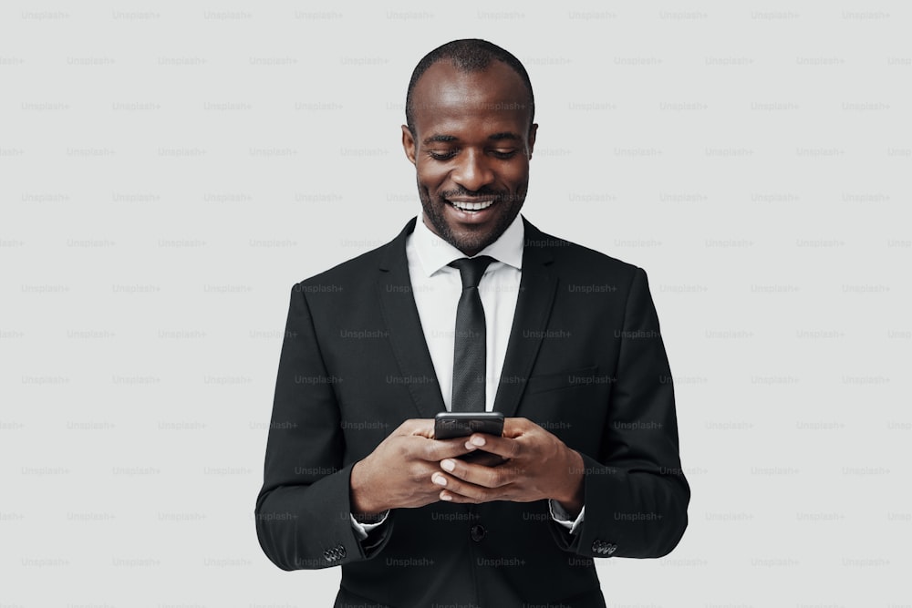 Jeune homme africain moderne en tenue de soirée à l’aide d’un téléphone intelligent et souriant tout en se tenant debout sur fond gris