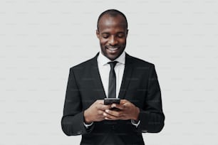 Modern young African man in formalwear using smart phone and smiling while standing against grey background