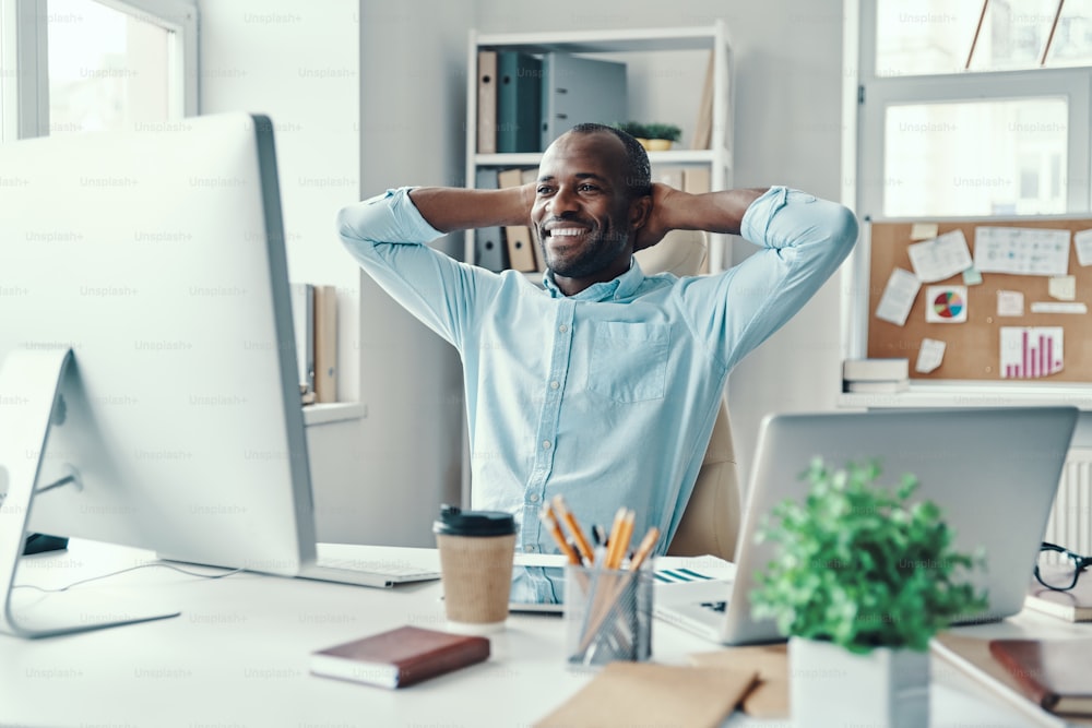 Jovem africano bonito de camisa mantendo as mãos atrás da cabeça e sorrindo enquanto trabalhava no escritório