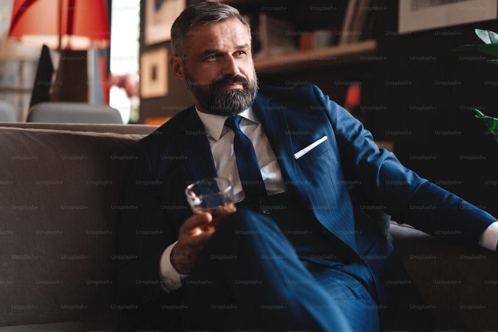 Hombre de negocios guapo y exitoso con un elegante traje que sostiene un whisky de vidrio mientras está sentado en el sofá de la oficina
