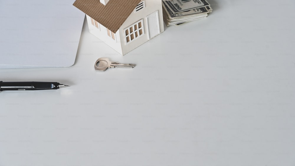 House model, rental agreement or house purchase agreement, money, paper, pen and the property's key on the white desk.