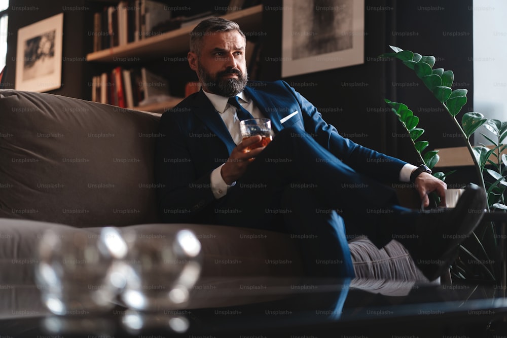 Handsome and successful businessman in stylish suit holding glass whiskey while sitting on the sofa at office