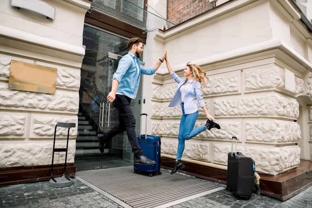Young cheerful couple with suitcases jumping and giving five, having fun, before arriving to the hotel. Couple having fun outdoors, near the old city building.