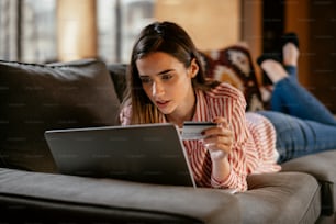 Junge Frau liegt mit Laptop auf dem Sofa und shoppt online