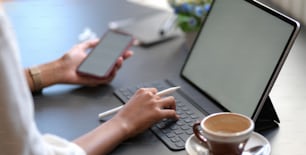 Vista de perto de uma jovem empresária digitando em um tablet digital enquanto usa um smartphone de tela em branco na sala de escritório