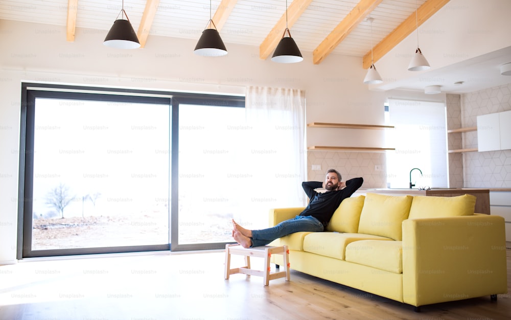 Homme mûr joyeux assis sur le canapé dans une maison non meublée, se déplaçant dans un nouveau concept de maison.
