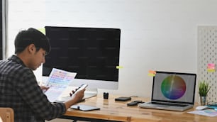 Redakteur sein aussehendes Smartphone und hält Farbpapier am Büroarbeitsplatz des Grafikdesigners.