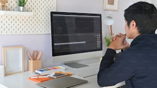 作業机でビデオ編集プログラムを使用している若い男性の後ろからのトリミングショットビュー。