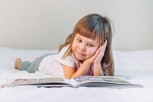 Beautiful little girl reading book lying in the bed.