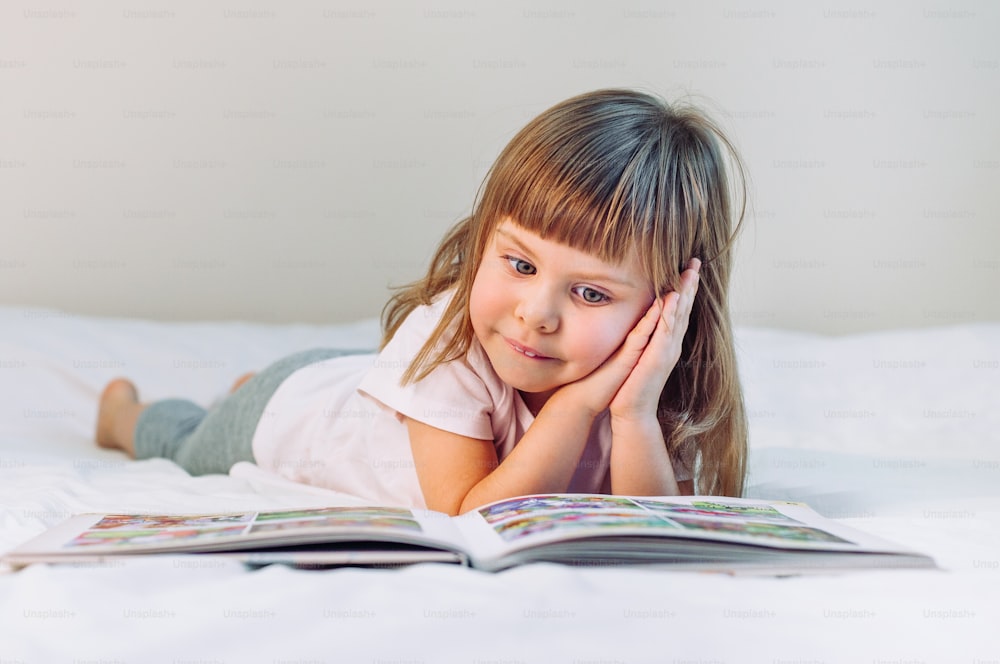 Beautiful little girl reading book lying in the bed.