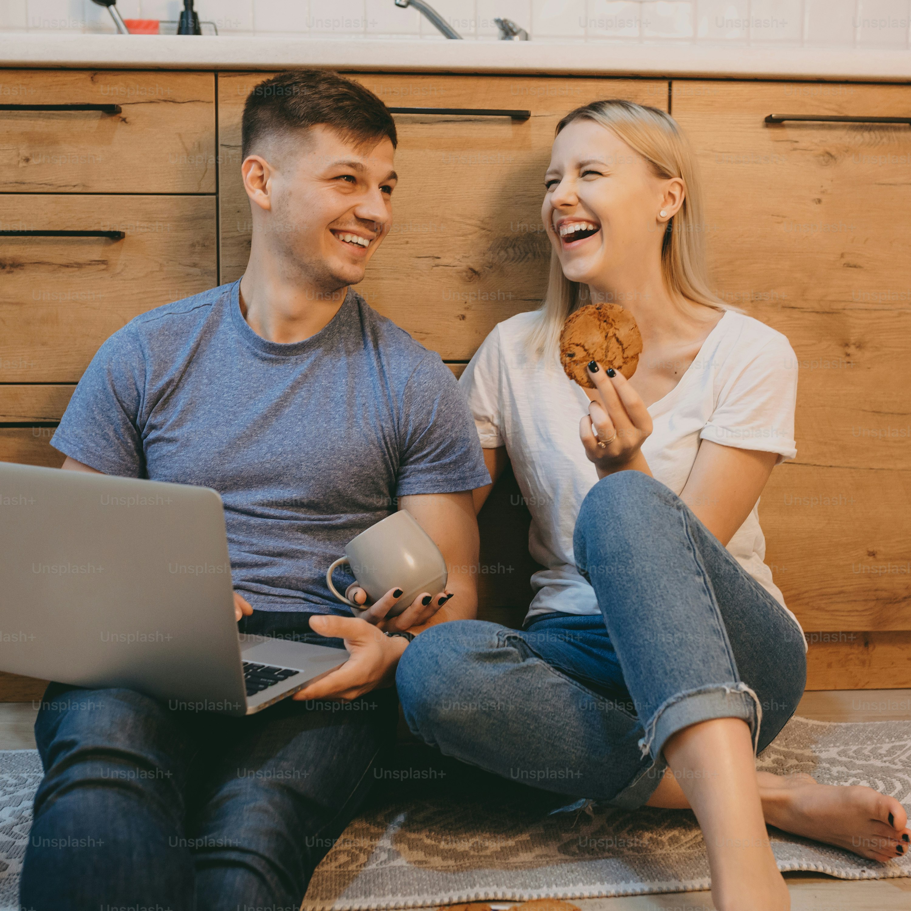 Charming young couple laughing while sitting on the floor in the kitchen in the morning while man is working at his laptop and woman is eating a cookie.