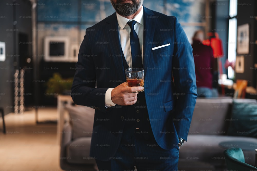 Mature handsome businessman stand and hold glass of whiskey in his own office