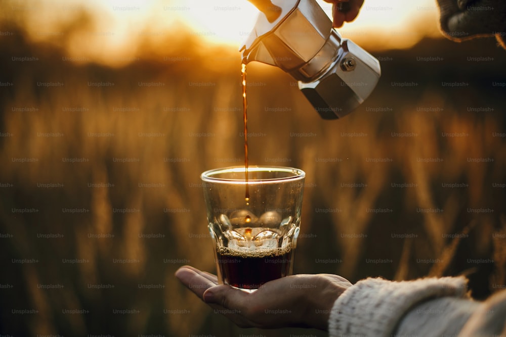Hipster che versa caffè caldo fresco dalla caffettiera geyser in una tazza di vetro in una luce calda e soleggiata nelle erbe della campagna rurale. Momento di tranquillità atmosferica. Preparazione alternativa del caffè in viaggio