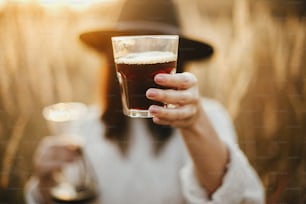 Hipster-Frau hält heißen Kaffee in Glastasse auf dem Hintergrund von ländlichen Kräutern im Sonnenuntergang. Alternative Kaffeezubereitung im Freien bei sonnigem Licht. Atmosphärischer rustikaler Moment. Reisen und Fernweh