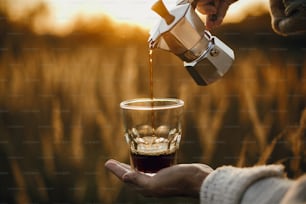 Voyageur versant du café chaud frais de la cafetière geyser dans une tasse en verre dans une lumière chaude et ensoleillée dans les herbes de la campagne rurale. Moment de tranquillité atmosphérique. Préparation alternative du café en voyage