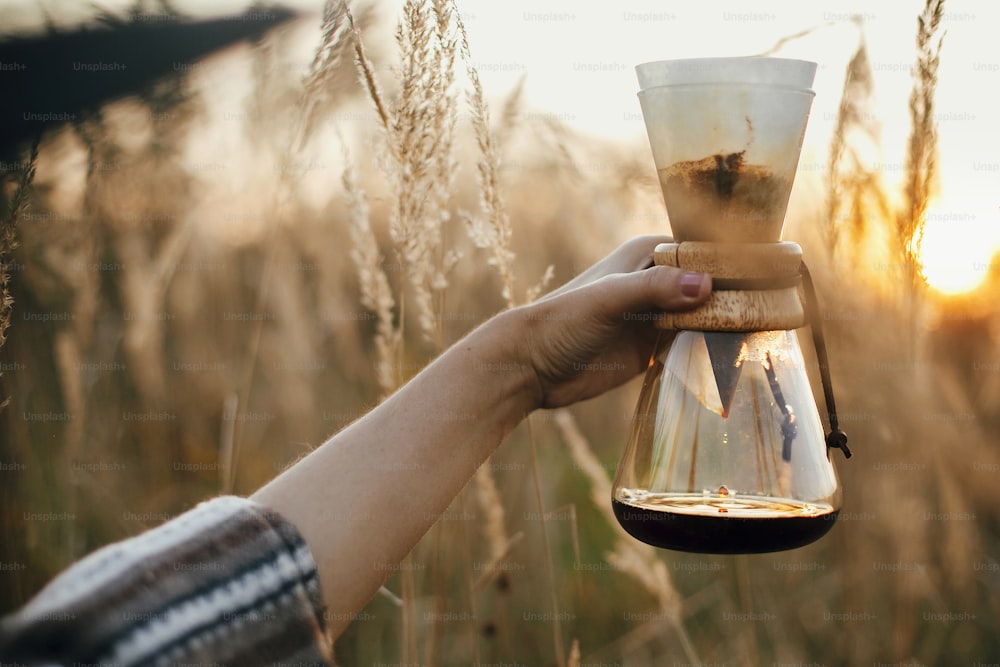 Préparation alternative du café à l’extérieur en voyage. Main tenant du café chaud dans une fiole en verre avec filtre sur fond de lumière chaude ensoleillée dans les herbes rurales. Moment tranquille rustique atmosphérique. Espace de copie