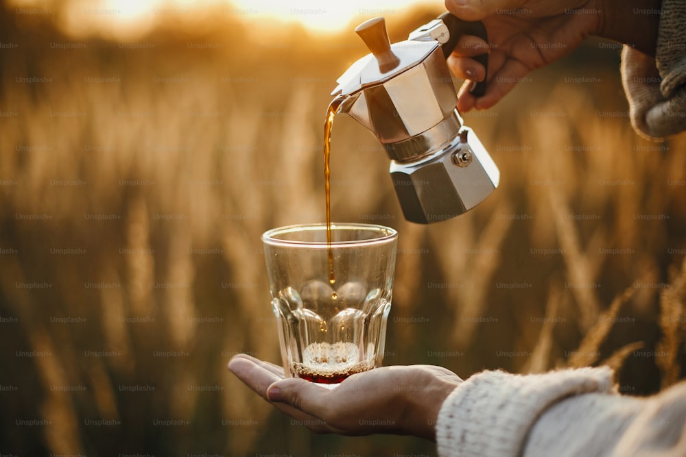 Preparación alternativa de café en viajes. Verter café caliente recién hecho de la cafetera del géiser en una taza de vidrio con una luz cálida y soleada en hierbas rurales del campo. Momento atmosférico. Imagen vertical
