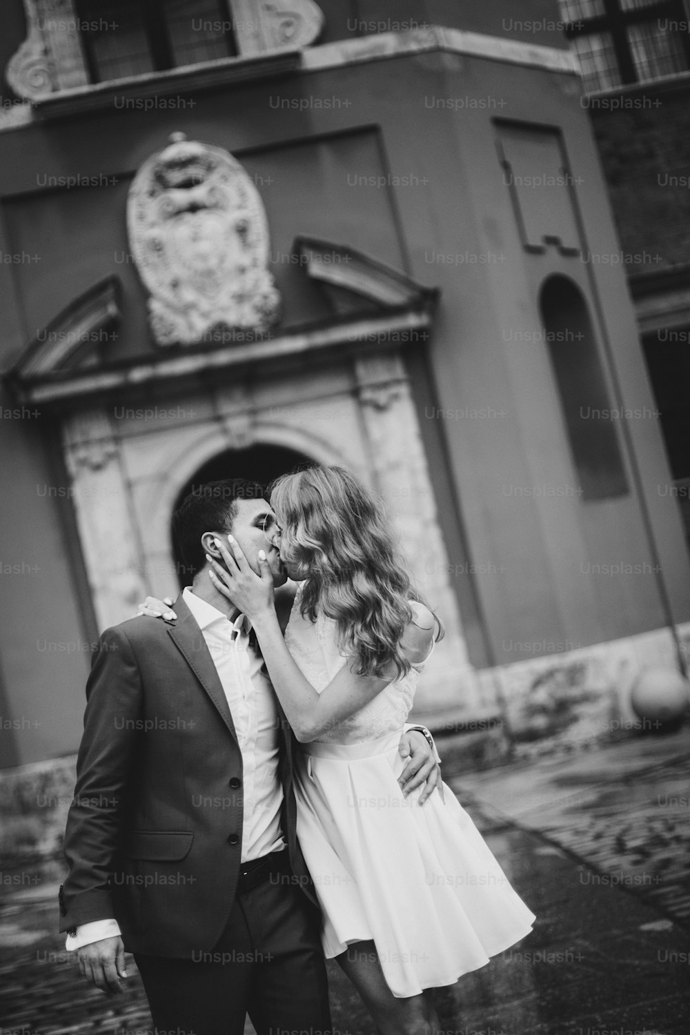 Stylish couple walking and kissing in european city street on background of old architecture. Fashionable bride and groom in love enjoying day, embracing in city. Traveling together in Europe