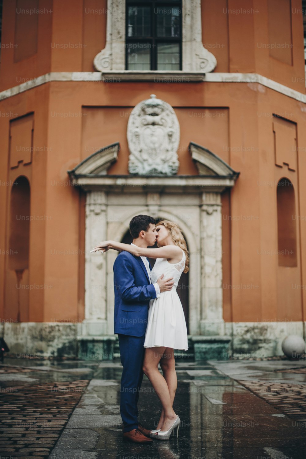 Coppia elegante che si bacia nella strada della città europea sullo sfondo della vecchia architettura. Sposa e sposo alla moda innamorati che si godono il giorno, abbracciandosi in città. Viaggiare insieme in Europa