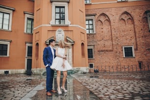 Stylish couple dancing in european city street on background of old architecture. Fashionable man and woman in love dancing with passion in city. Traveling together in Europe