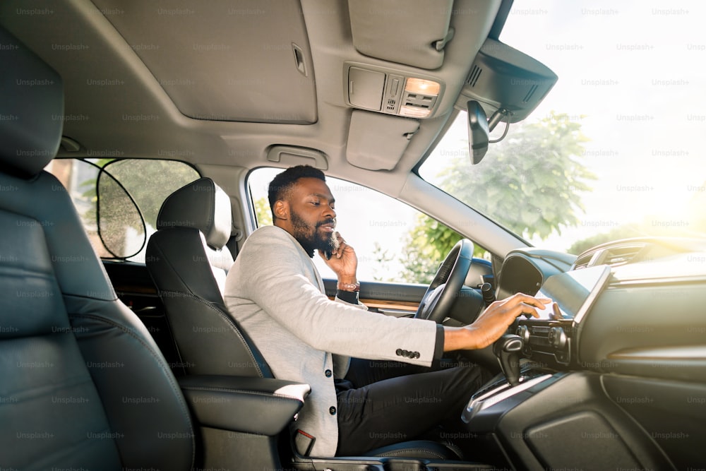 Hombre de negocios africano hablando por teléfono móvil dentro de un coche y tocando la tableta. Joven emprendedor trabajando durante el viaje a la oficina en un coche de lujo