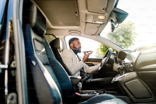 Young successful African American businessman talking on speakerphone through microphone with client, sitting in the expensive car. Negotiations and business meetings