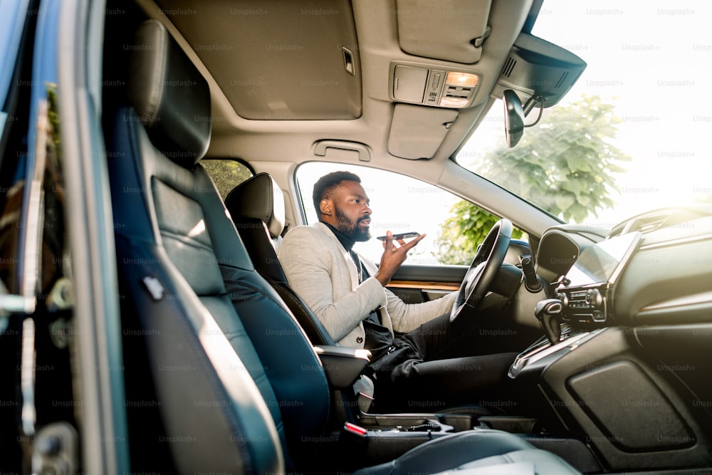 Junger erfolgreicher afroamerikanischer Geschäftsmann, der über die Freisprecheinrichtung über das Mikrofon mit dem Kunden spricht und im teuren Auto sitzt. Verhandlungen und Geschäftstreffen