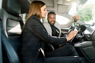 Handsome cheerful young african man holding car keys from his new luxury car, while sitting in the car together with pretty woman car salesmanager holding a tablet.