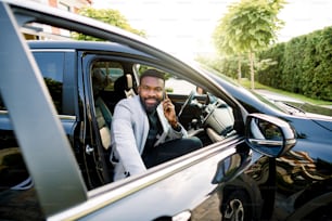 Happy smiling African american businessman in smart casual suit speaking on smartphone while getting in the black car on passenger seat