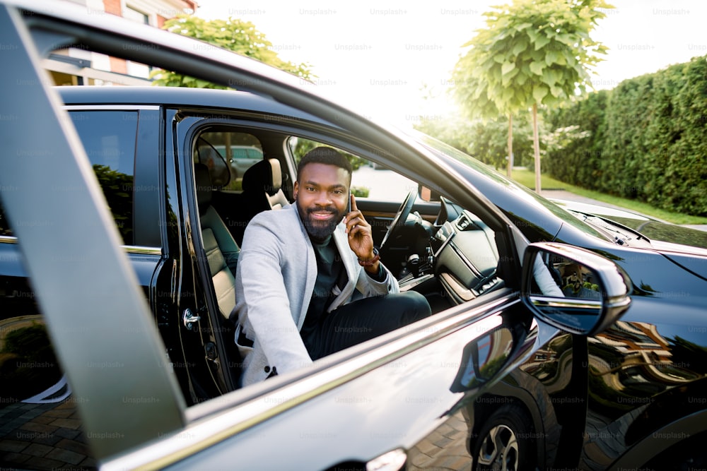 Heureux homme d’affaires afro-américain souriant dans un costume décontracté élégant parlant sur un smartphone tout en montant dans la voiture noire sur le siège passager