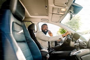 Young African American man in smart casual business wear, using phone sitting in the car, side view.