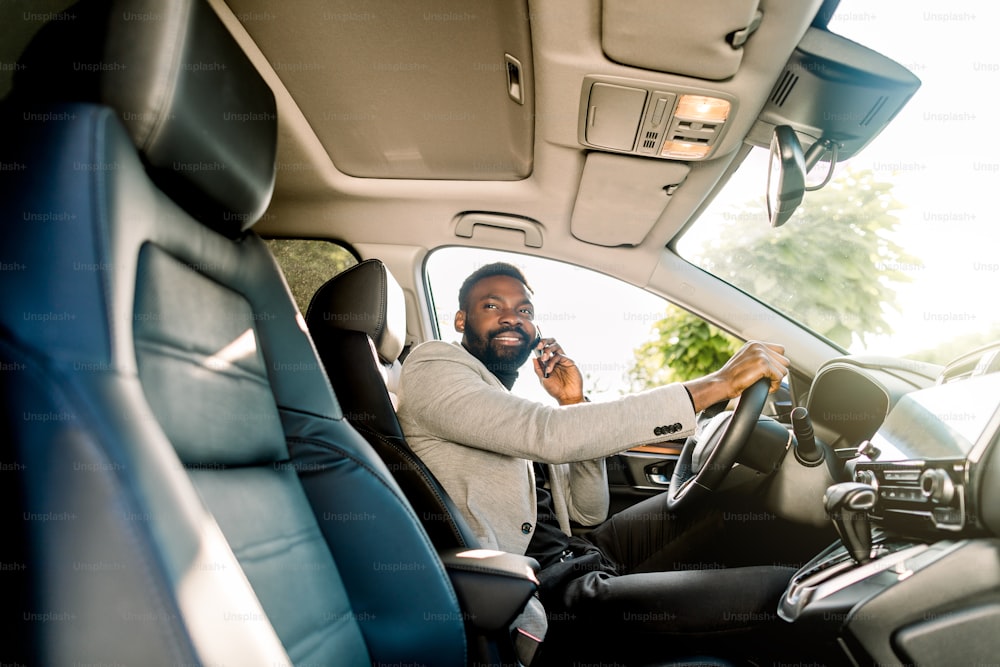 Joven hombre afroamericano en ropa de negocios casual elegante, usando el teléfono sentado en el automóvil, vista lateral.