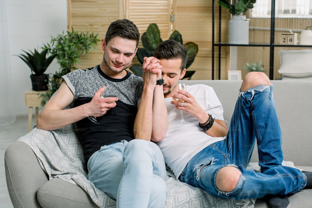 Young gay men holding hands on sofa at home. Shy gay couple showing love and romance, drinking wine