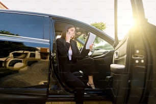 Giovane donna d'affari seria vestita in abiti formali che parla con il partner tramite telefono cellulare e utilizza appunti mentre è seduta in un'automobile nera di lusso.