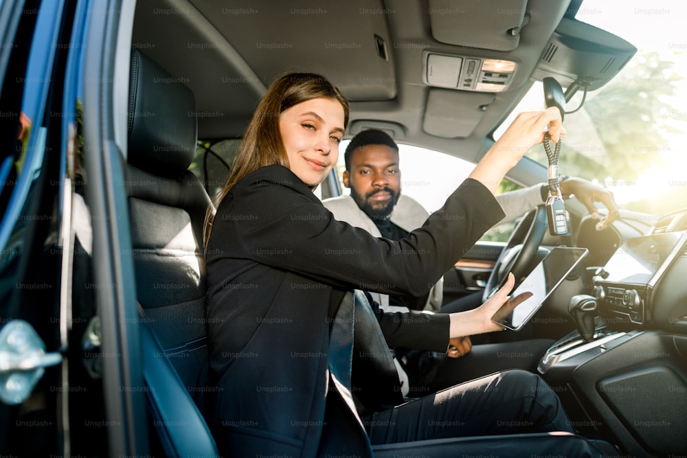 Young pretty Caucasian woman car dealer holding car key and digital tablet and looking at camera, while sitting in the car with her smiling handsome client, African businessman.