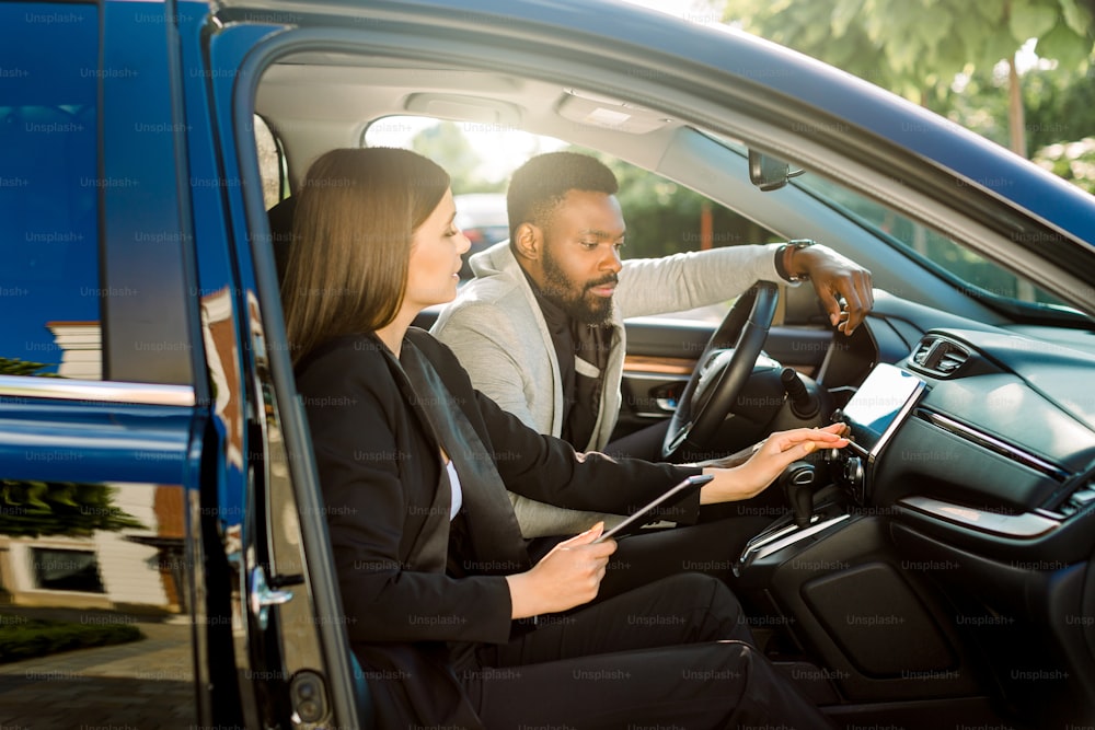 Two smiling business people, Caucasian woman and African man sitting together in the car, using digital tablet and working. Discuss business strategy while driving to a meeting.