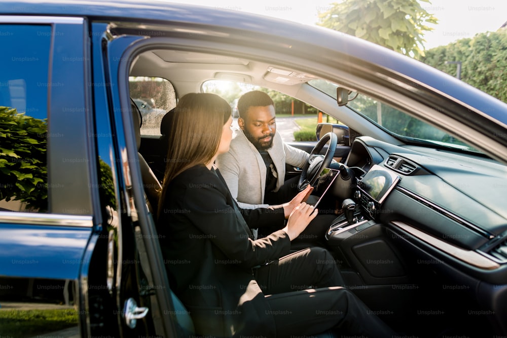 Giovane uomo afroamericano che comunica con una collega caucasica durante la guida in auto alla riunione di lavoro che discute idee di avvio e presentazione da tablet digitale.