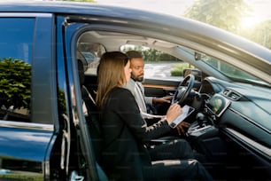 due uomini d'affari felici, uomo africano e donna caucasica, utilizzando la tavoletta digitale dei dispositivi elettronici mentre si siede nell'auto nera.