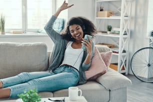 Playful young African woman in casual clothing listening music and smiling while resting at home