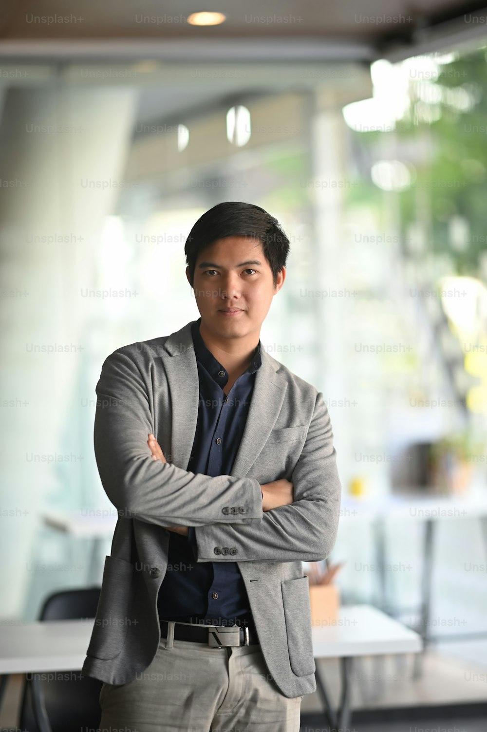 Foto de um jovem empresário confiante em terno cinza mantendo os braços cruzados enquanto estava ao lado da mesa de trabalho.