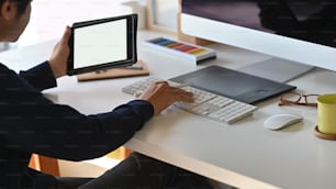 Cropped shot of young stylish producer holding on white blank screen tablet and typing on keyboard over the modern working desk background.