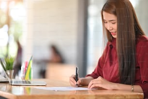 Portrait of beautiful woman looking good in red shirt while writing on the paper and sitting at the wooden working desk.