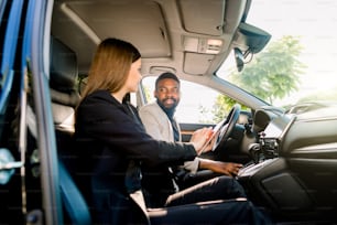 Beautiful business woman in black suit is using a digital tablet and smiling while sitting in the car together with her business partner, Handsome African man.