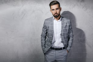 Handsome bearded man wearing gray checkered suit is posing against concrete wall