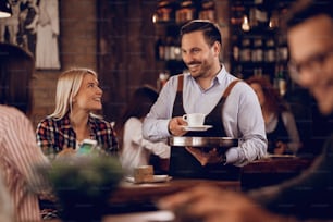Young happy waiter serving coffee while talking to female guest in a cafe.