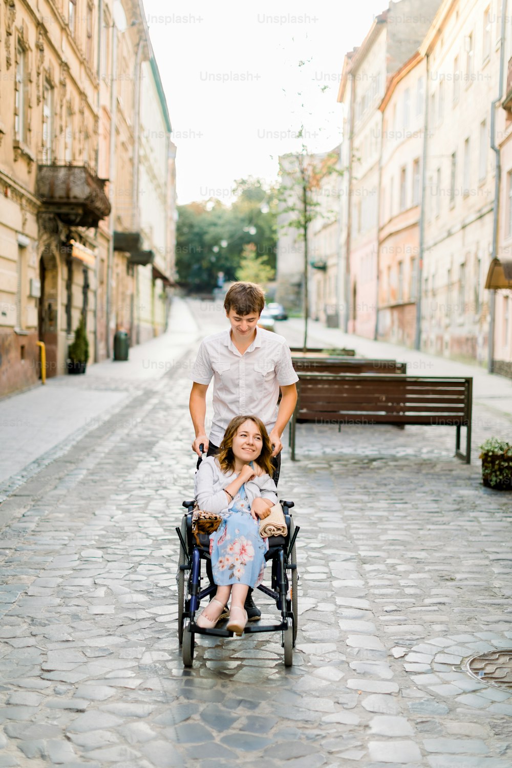 Young Couple In Wheelchair Strolling In The City. Pretty young woman in wheelchair and her boyfriend in the center of old city.