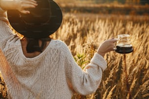 Hipster woman in hat holding hot coffee in glass kettle on background of rural herbs in sunset warm light. Alternative coffee brewing outdoors. Atmospheric rustic moment. Travel and Wanderlust