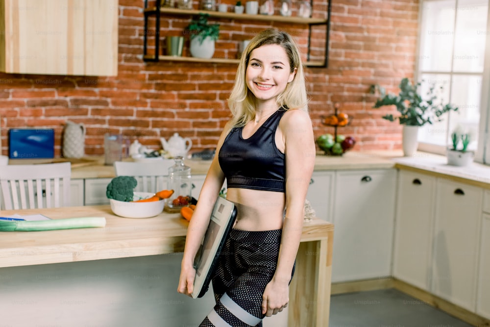 Close-up portrait of beautiful fit woman standing in her kitchen with a weight scale in her hand on the background of brick wall ant kitchen table. Loosing weight, diet, and healthy nutrition