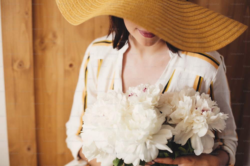 Portrait of boho woman in hat holding white peony bouquet on rustic wooden background. Stylish hipster girl in dress posing with peonies. Happy Mothers day. International Womens days