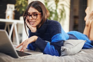 Jolie brune caucasienne en pull bleu et noir allongée sur le ventre dans le lit et tapant sur un ordinateur portable.